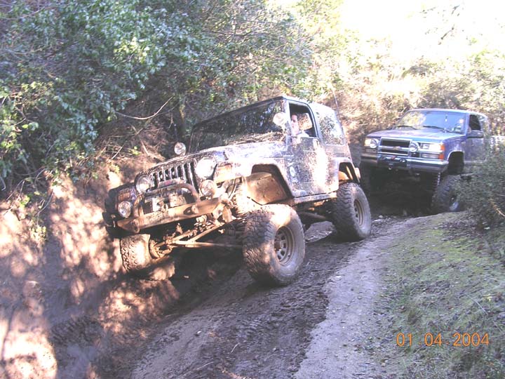 Mud-covered topless Jeep Wrangler leaning in a rut with a lifted Chevy pickup behind