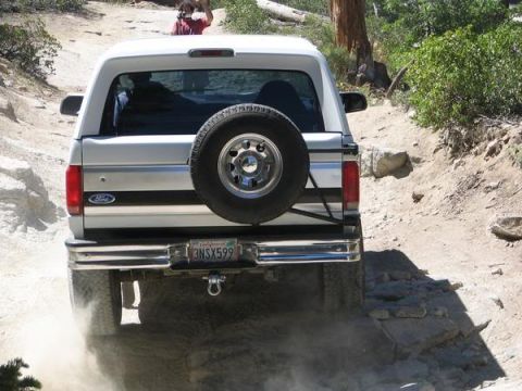 1995 Ford Bronco - A white Ford Bronco navigates a rocky, off-road trail, kicking up dust.