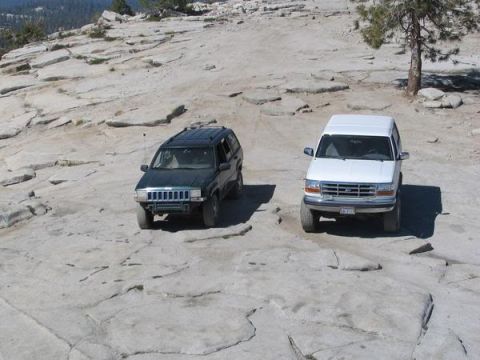1995 Ford Bronco - Two SUVs are parked on a large, flat rock formation with sparse trees and distant hills in the background.