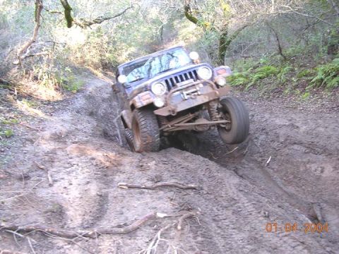 1994 Ford Bronco - A mud-covered Jeep Wrangler crawls at a steep angle down a rutted clay trail in the woods, flexing its solid front axle with one wheel droo…