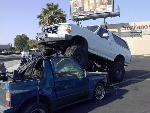 1995 Ford Bronco - A white 1995 Ford Bronco is parked on top of a severely damaged blue pickup truck.
