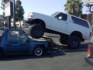 1995 Ford Bronco - A white Ford Bronco is perched atop a severely damaged blue car, indicating a recent collision.