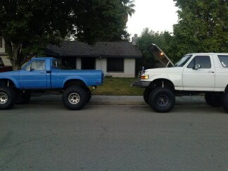 1995 Ford Bronco - Two heavily modified trucks, a white Ford Bronco and a blue pickup, are parked side-by-side on a residential street.
