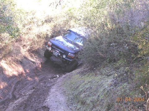 1994 Ford Bronco - Looking down from above at a dark blue full-size Ford Bronco working up a narrow, rutted dirt trail through brush, with deep tire ruts in t…