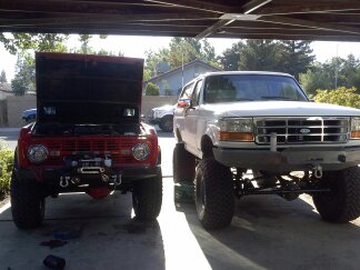 1995 Ford Bronco - Two lifted Ford Broncos, one red with its hood up and a winch, the other white, are parked side-by-side.