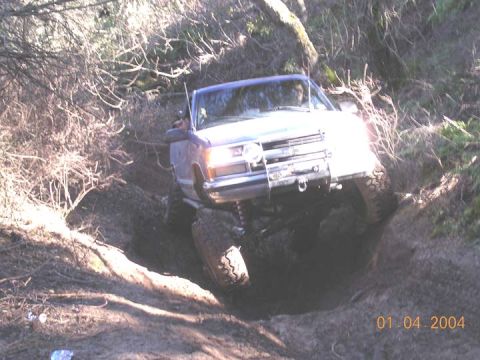 1994 Ford Bronco - A lifted two-tone blue and white Ford full-size 4x4 crawls through a muddy rutted hole on a wooded trail, articulating hard with the driver…