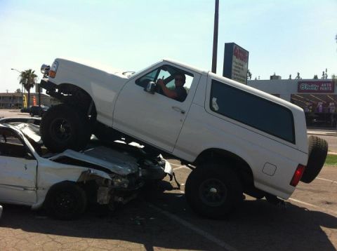 1995 Ford Bronco - A white Ford Bronco is parked on top of a severely damaged white car, with the driver giving a thumbs-up.