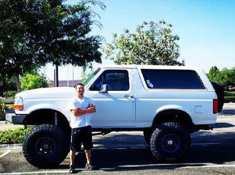 1995 Ford Bronco - A man stands beside a white 1995 Ford Bronco, which features a lifted suspension and oversized tires.