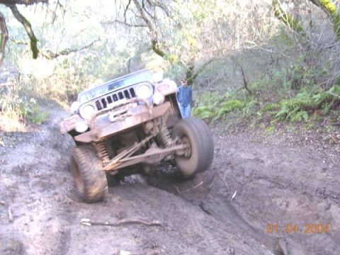 1994 Ford Bronco - A lifted 4x4 climbs out of a deep mud rut on a wooded trail, front end raised high so the coil-sprung solid front axle, radius arms and ste…