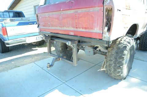 1979 Ford Bronco - The rear of a red Ford Bronco shows a custom-built hitch and large tires, with a blue pickup truck parked nearby.