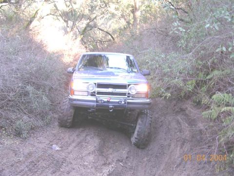 1994 Ford Bronco - A lifted silver-blue Ford Bronco climbs a rutted dirt trail cut through brushy woods, shot head-on with its headlights on and front axle ar…
