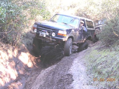 1994 Ford Bronco - A mud-splattered lifted full-size Ford Bronco on oversized mud tires crawls out of a deeply rutted trail gully, with a bumper-mounted winch…