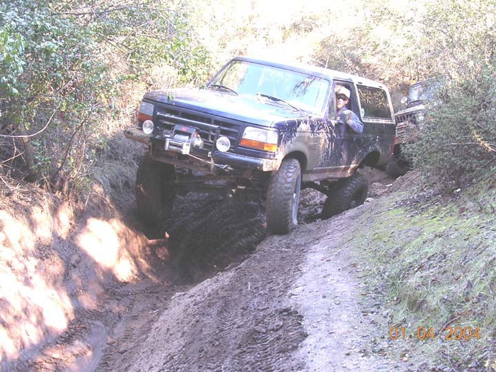 Muddy lifted Ford Bronco with front winch climbing a rutted dirt trail gully