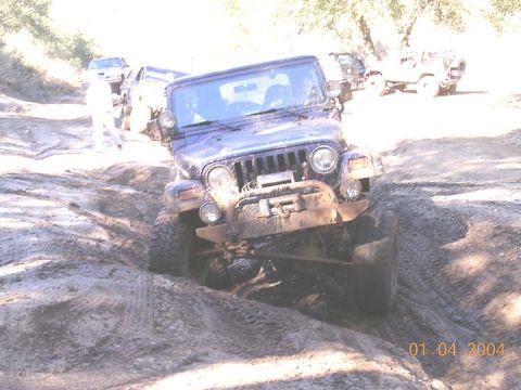 1994 Ford Bronco - A mud-covered dark Jeep Wrangler TJ with round headlights, a seven-slot grille, tube bumper and winch drops its front wheels into a deep ru…