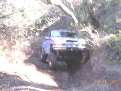 1994 Ford Bronco - Blurry action shot of a lifted light-colored 4x4 pickup dropping into a steep dirt gully on a brushy hillside trail, front wheels articulat…