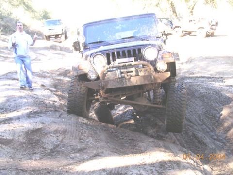 1994 Ford Bronco - A mud-splattered blue lifted Jeep Wrangler TJ articulating through a rutted rock-and-dirt trail, front view with a winch-equipped bumper, e…