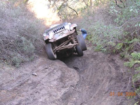 1994 Ford Bronco - Light-colored open-top Jeep Wrangler lifting a front wheel as it climbs a rutted dirt trail through wooded terrain, exposing the front axle…