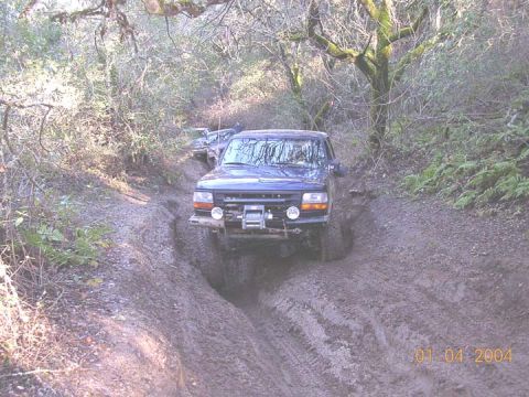 1994 Ford Bronco - Dark blue lifted Ford 4x4 crawling through a deeply rutted muddy trail in a mossy wooded forest, seen head-on from downhill. It runs oversi…