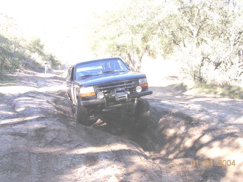 1994 Ford Bronco - Dark blue lifted Ford Bronco crawling a rutted dirt trail, front end dropped into a deep washout, with a Warn winch mounted in a tube front…