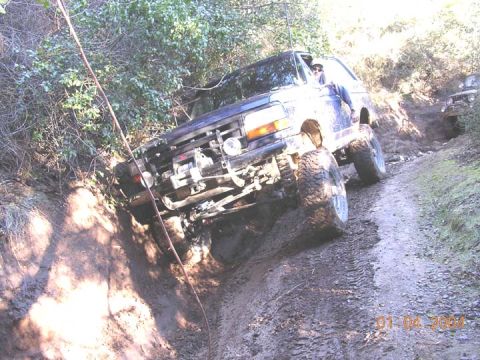 1994 Ford Bronco - A lifted white Ford Bronco on oversized mud-terrain tires tips steeply off-camber in a muddy rutted trail, front bumper cut away to expose…