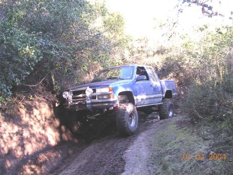 1994 Ford Bronco - A mud-splattered blue Chevrolet C/K extended-cab pickup on a lifted suspension with oversized mud-terrain tires climbs off-camber onto a ru…