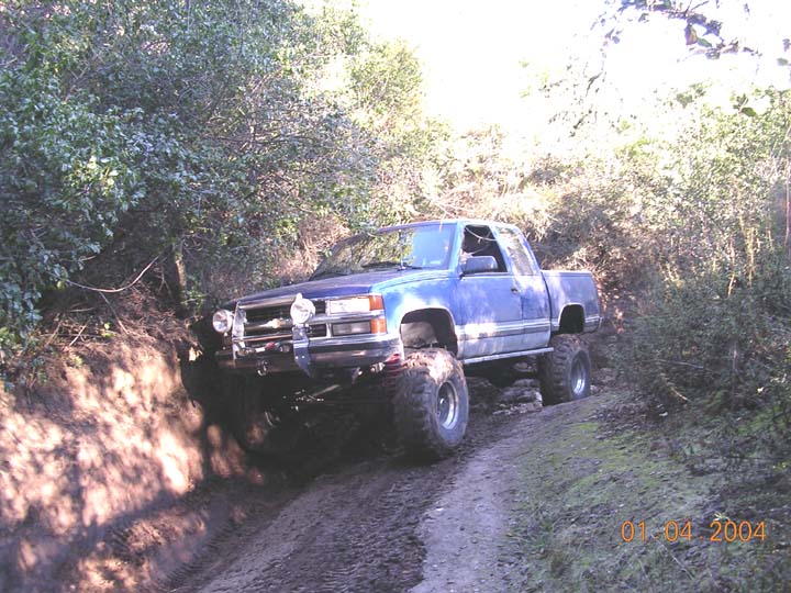Lifted blue Chevy extended-cab pickup with winch bumper on a muddy wooded trail