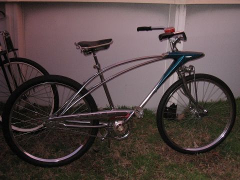 Chrome cruiser bicycle with teal front fairing and wide rear tire. Another bicycle is partially visible behind