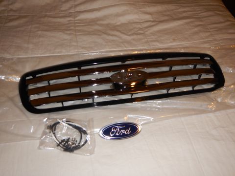 2003 Ford Crown Victoria - black-and-chrome front grille beside a plastic hardware bag and a blue Ford oval emblem on a white sheet.