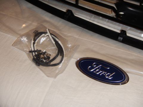 2003 Ford Crown Victoria - bag of grille screws and clips beside a glossy blue Ford oval emblem, with a grille edge in the background.