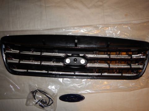 2003 Ford Crown Victoria - black plastic front grille with chrome horizontal bars and a Ford blue oval emblem, shown with a bagged wiring harness and a separate Ford…