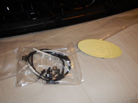 2003 Ford Crown Victoria - clear plastic bag holding a wiring harness and connectors beside a primer-yellow blank Ford oval badge, with a dark grille edge above.