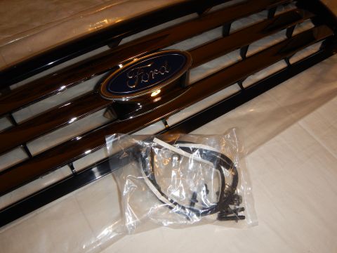2003 Ford Crown Victoria - Angled close-up of a black Ford grille's chrome trim and blue oval emblem, with a bagged wiring harness and screws resting in front.