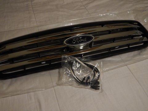 2003 Ford Crown Victoria - Three-quarter view of a black Ford grille with chrome bar accents and a blue oval emblem, displayed beside a bagged wiring harness.
