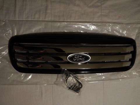 2003 Ford Crown Victoria - glossy black Ford front grille with chrome bars and a centered blue oval emblem, laid on plastic wrap with a small wiring harness.