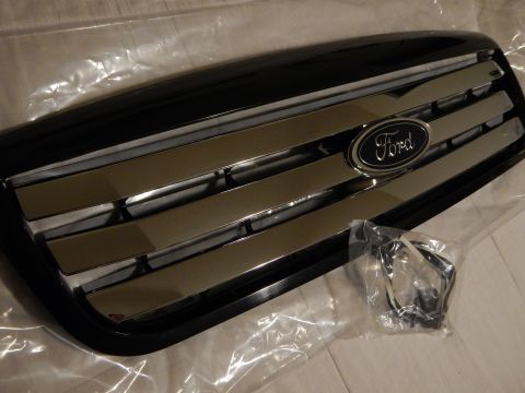 2003 Ford Crown Victoria - Angled view of a black Ford grille with chrome horizontal bars and a blue oval emblem, shown with a bagged wiring harness.