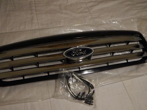 2003 Ford Crown Victoria - Three-quarter view of a black Ford grille with chrome bar accents and a centered blue oval emblem, beside a bagged harness and screws.