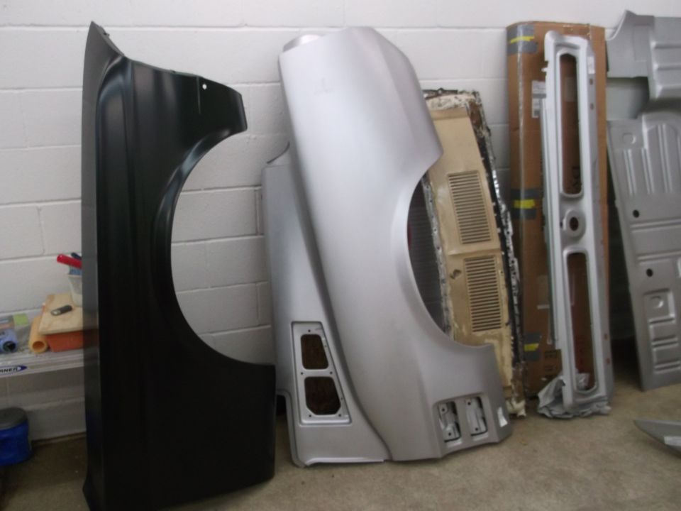 Two car fenders, one black and one silver, leaning against a brick wall with other car body parts behind them