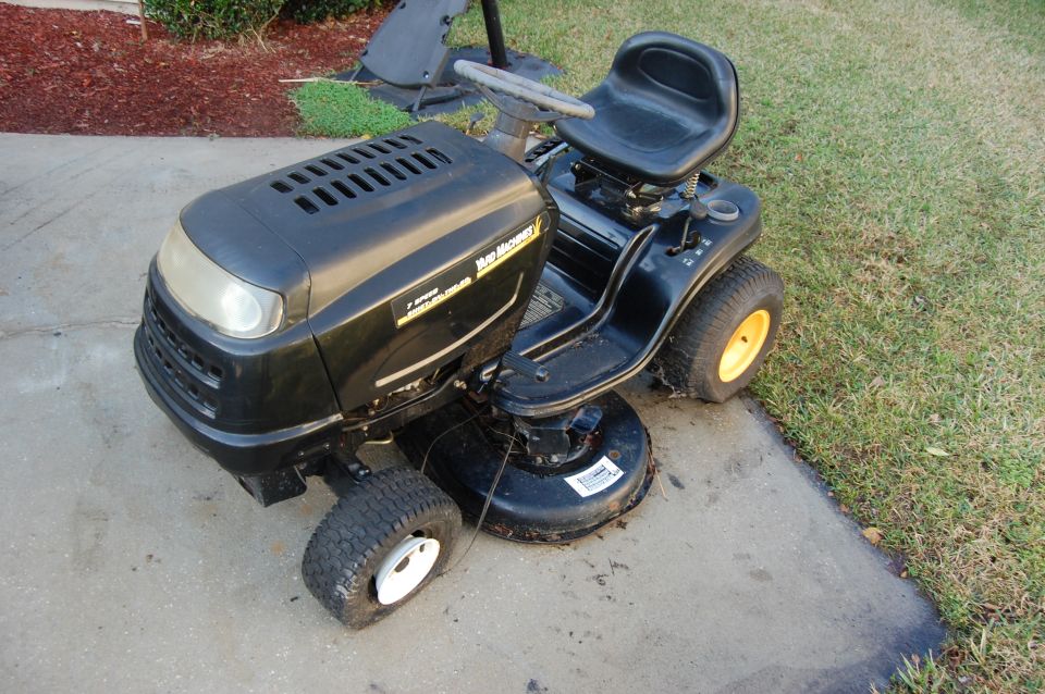 Black riding lawn mower parked on concrete and grass. It has a yellow-rimmed rear wheel and a white-rimmed front wheel