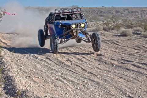 1996 Ford Bronco - A blue off-road buggy kicks up dust as it jumps over a dirt track in a desert landscape.