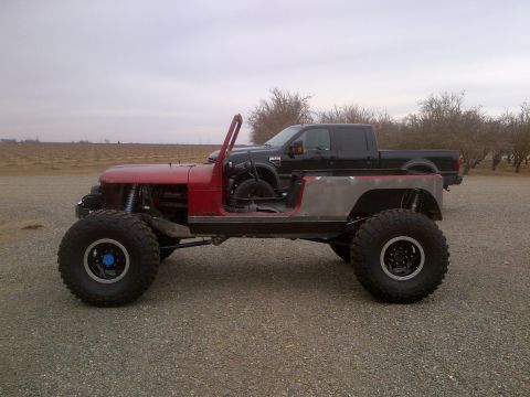 1985 Jeep CJ7 - A heavily modified Jeep-style vehicle sits beside a black pickup truck under an overcast sky.