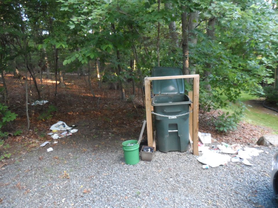 Trash cans and scattered debris in a wooded area with a gravel ground