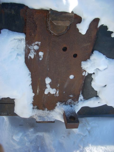 1963 Hummer H3 - A close-up view of a rusted metal bracket covered in snow, showing two circular holes and a rectangular attachment point.