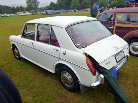 1969 MINI Cooper - A white sedan is parked on a grassy field with its trunk open, revealing items inside.