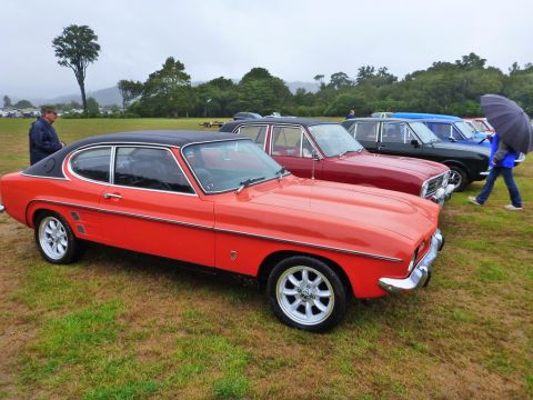 1969 MINI Cooper - An orange Ford Capri is parked in a line of cars on a grassy field under an overcast sky.