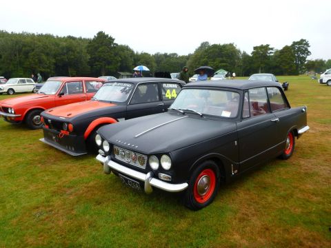 1969 MINI Cooper - A line of classic cars are parked on a grassy field under an overcast sky.