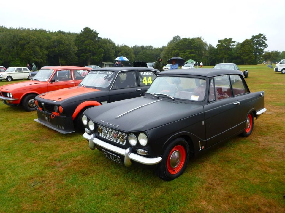 Black Triumph Herald with red wheels parked on grass next to other cars
