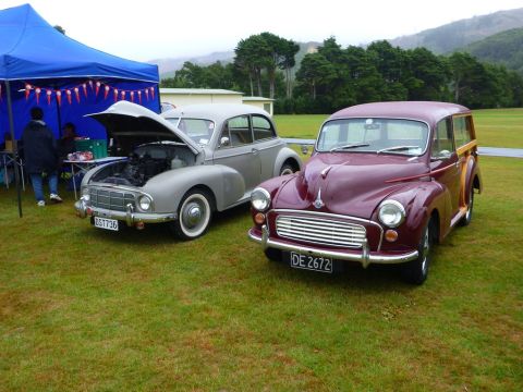 1969 MINI Cooper - Two vintage cars are displayed outdoors under a blue tent, with trees and hills in the distance.
