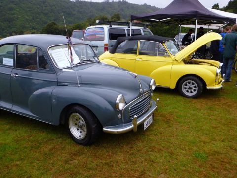 1969 MINI Cooper - Two classic British cars, a gray Morris Minor 1000 and a yellow convertible, are displayed on a grassy field.
