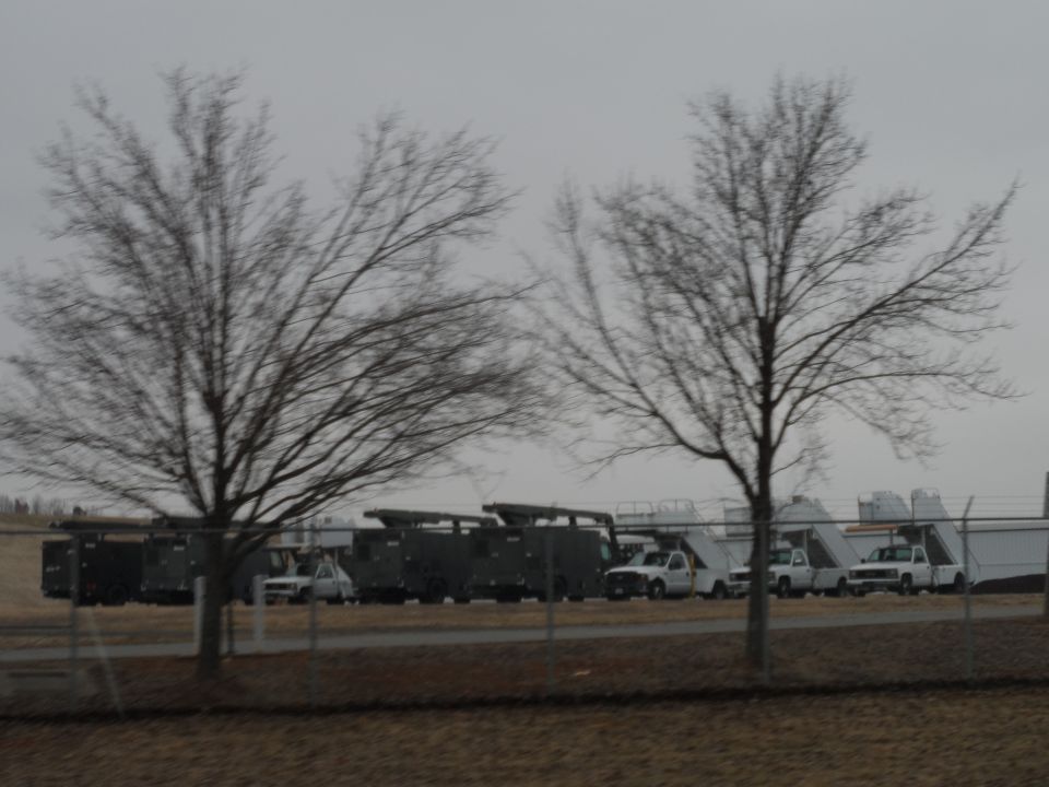Several white pickup trucks and dark utility vehicles parked in a lot with bare trees in the foreground