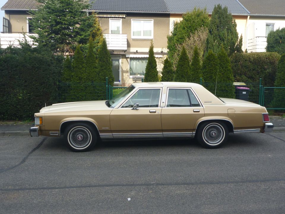 Side view of a gold two-tone 1984 Mercury Grand Marquis sedan parked on a street in front of hedges and houses